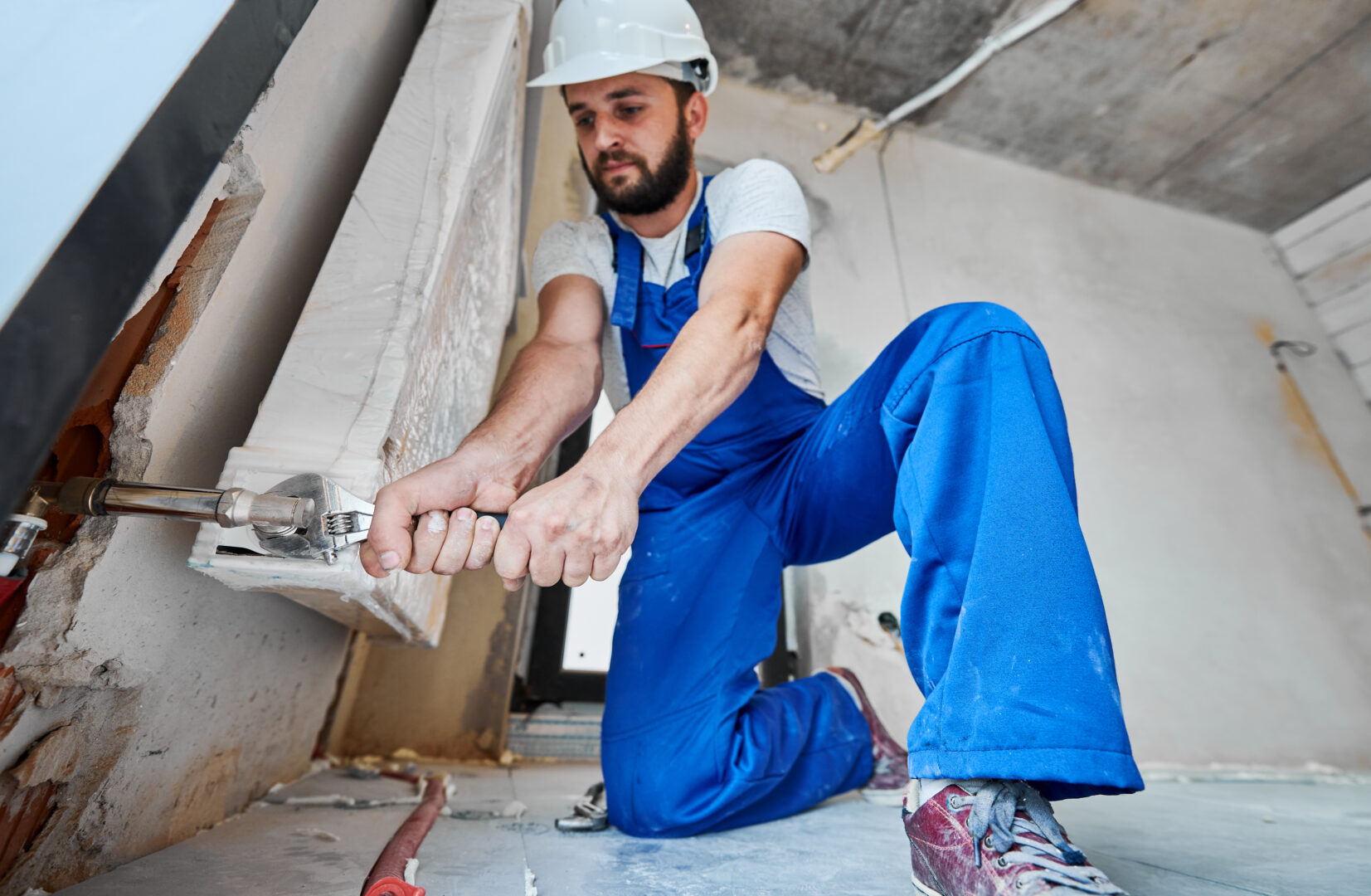 Horizontal snapshot of plumber screwing plumbing fittings with a wrench. Low angle view on man, wearing blue overalls and helmet, working with construction tool with both hands applying force